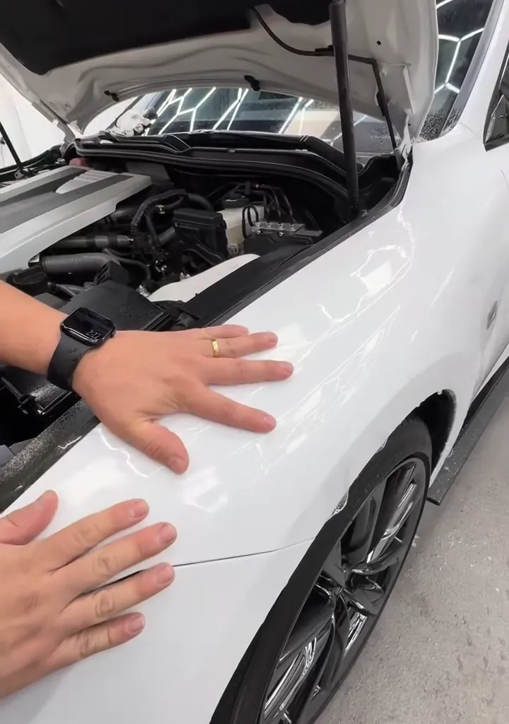 Hands smoothing clear film over a white hood, hexagon lights reflected in the paint
